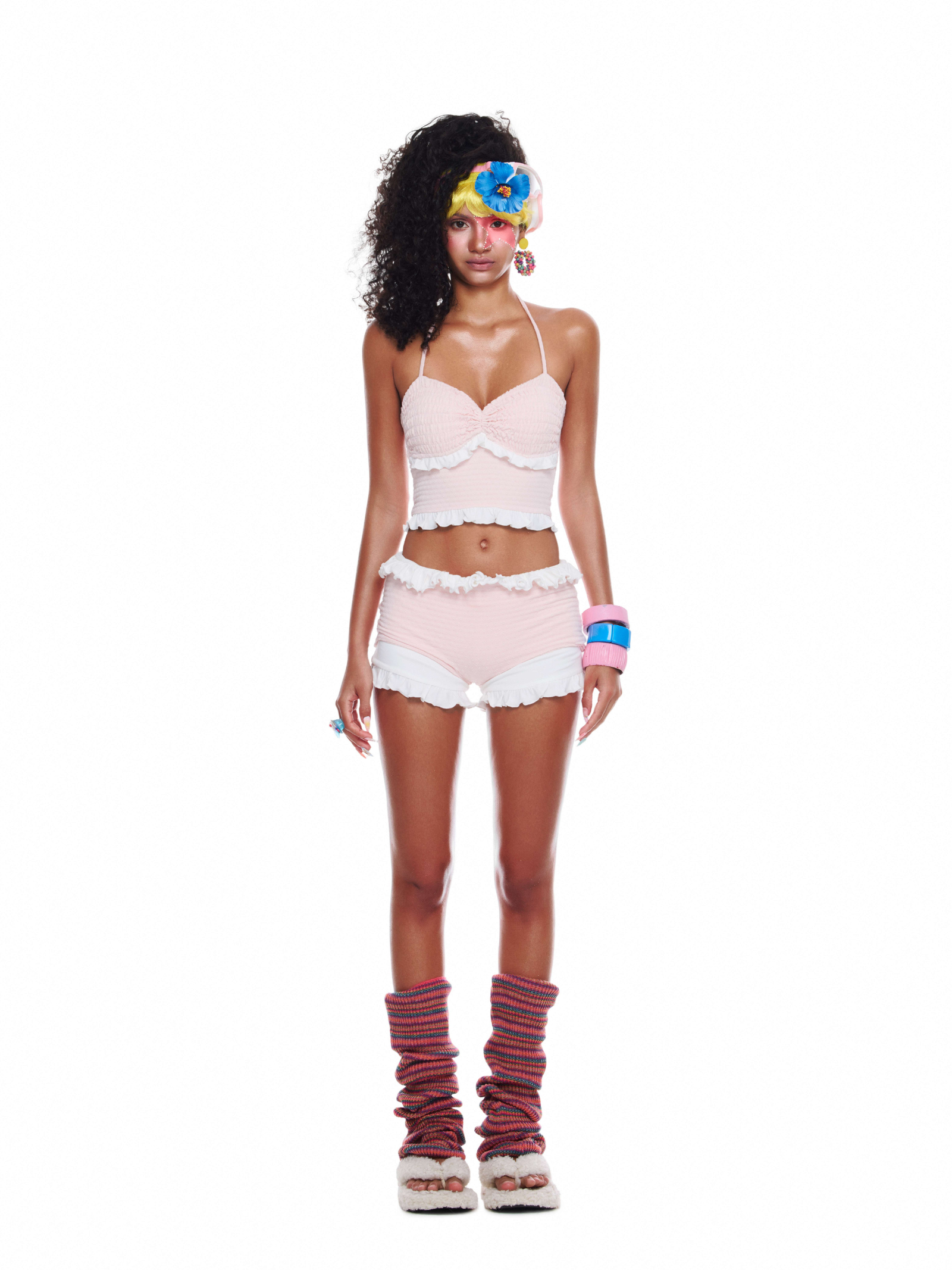 Model wearing a pastel pink beach outfit with knit crop top and ruffled shorts, styled with leg warmers and soft platform sandals.
