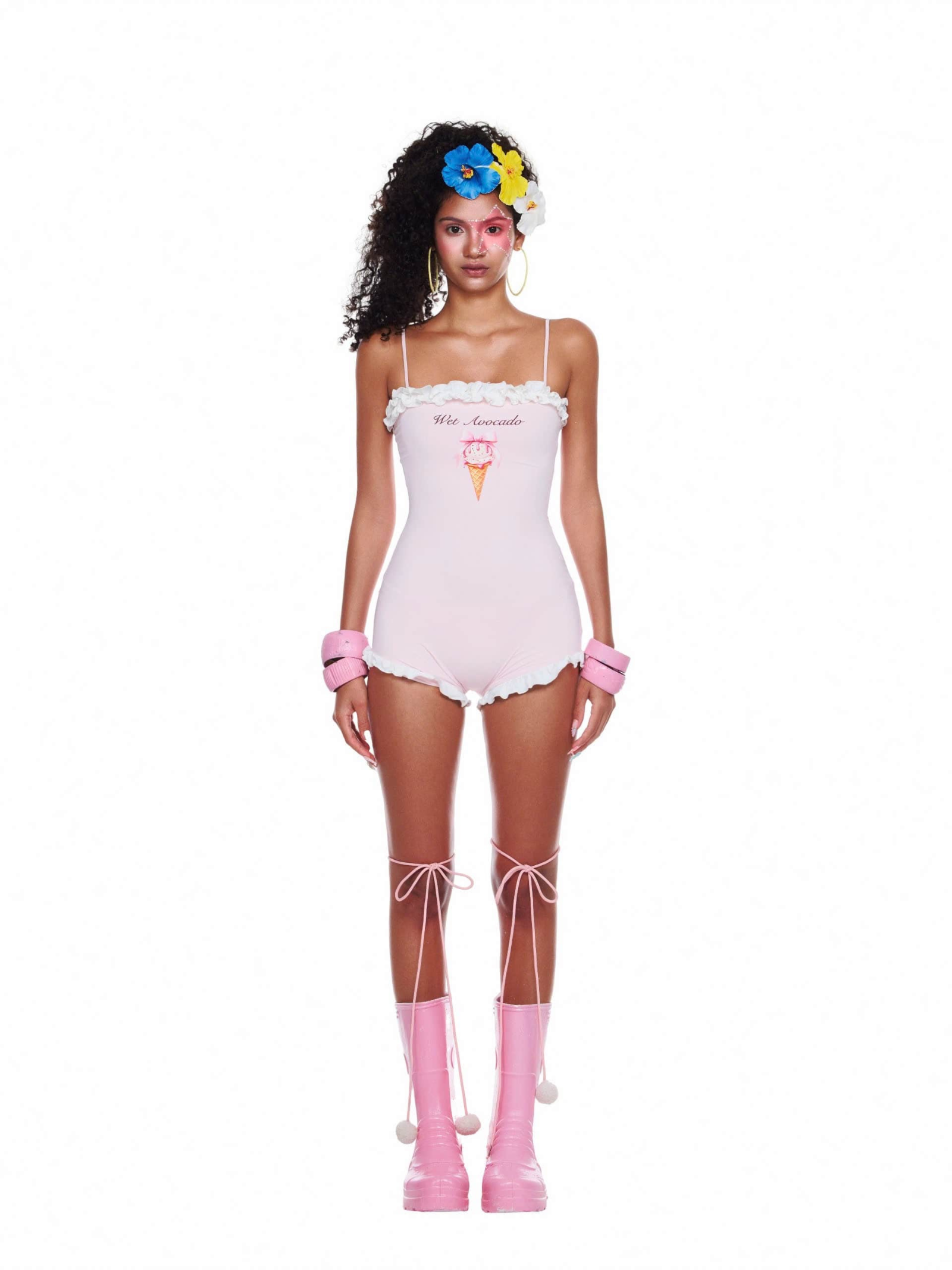 Model wearing a pastel pink beachwear swimsuit with ruffled trim, spaghetti straps, and printed ice cream graphic, styled with pink boots and accessories.
